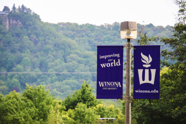 Purple banners with the mission statement hang from light poles across the WInona campus with a view of the bluffs in the background.