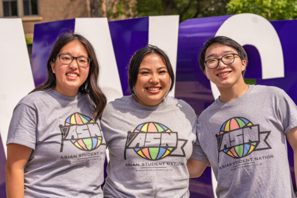 Three students wearing shirts from the Asian Student Nation club pose smiling on the WSU campus.