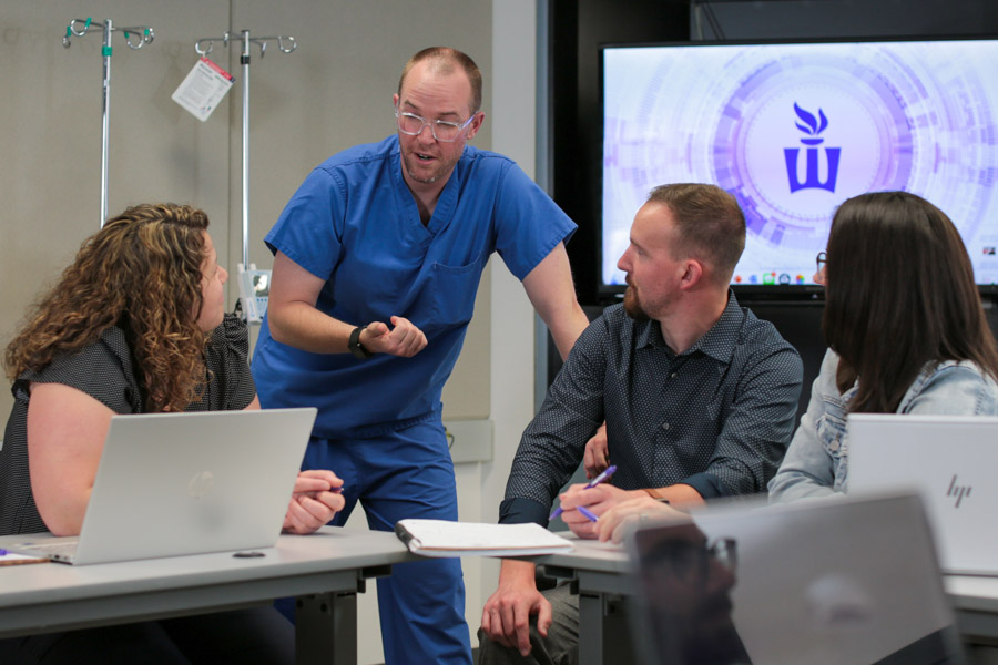 A group of healthcare professionals, one wearing scrubs, talk to one another.