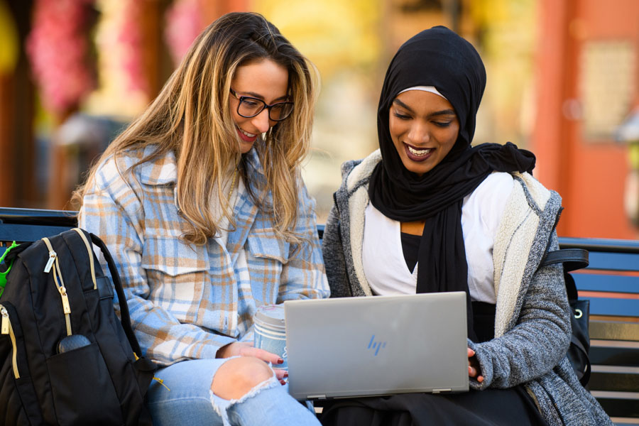 Two female students sit on a bench outside and look at a laptop together.