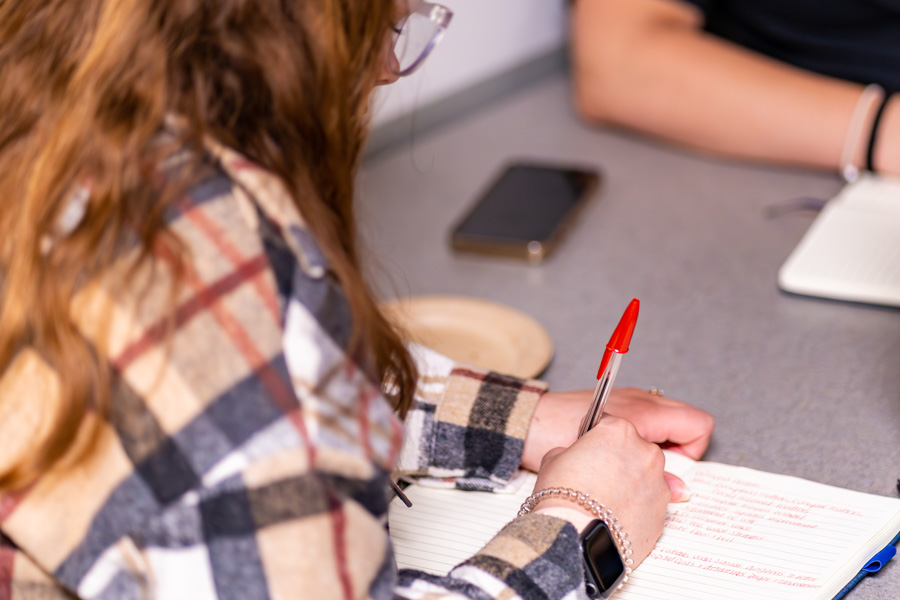 A student wearing glasses writes with a red pen in a notebook.