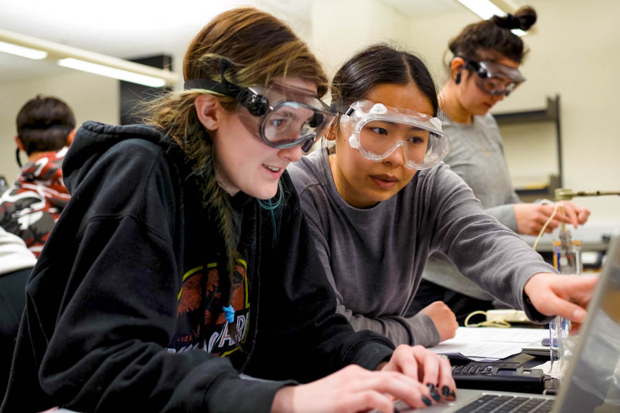 WSU students sitting at a table, looking at a laptop, and wearing lab goggles.