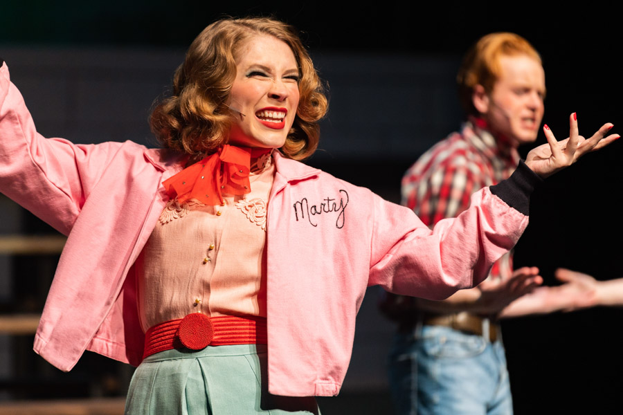 Female student wearing pink jacket performing in "Grease" production.