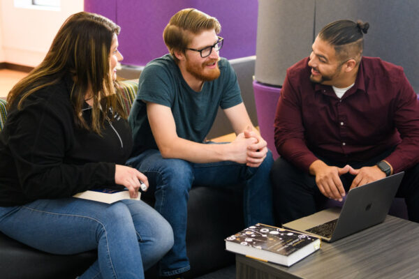 Three students sitting on a couch with a laptop and textbook in front of them.