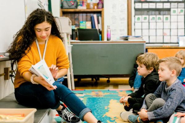 A female student reads to a group of kids in a classroom.