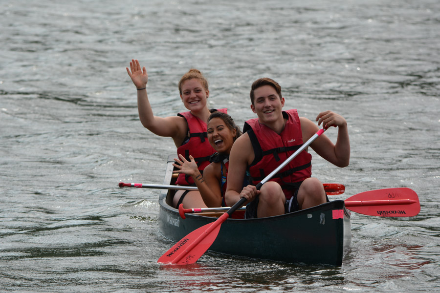 Three students wave and smile while they canoe together on Lake Winona.