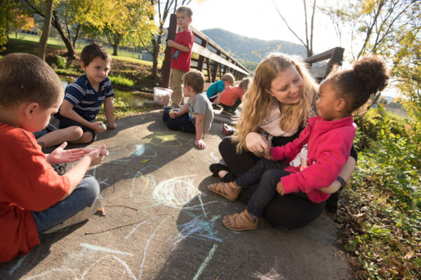 A female student draws in chalk with toddlers on a wooden bridge.