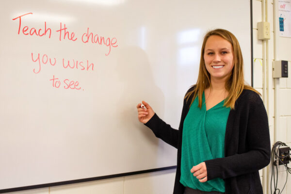 A teacher smiles in front of a classroom whiteboard that has