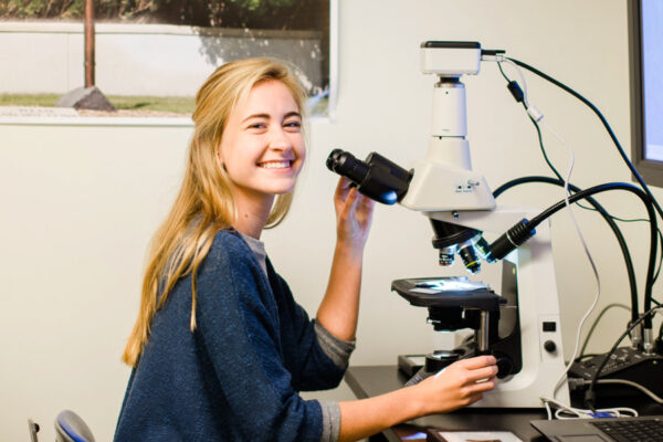A student looks up from a microscope and smiles.