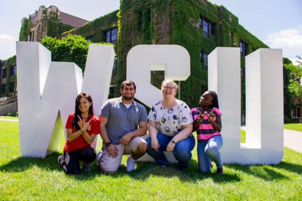 A group of students pose smiling in front of WSU letters on the Winona campus.