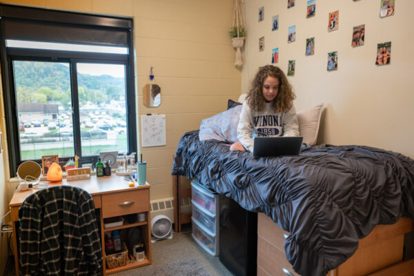 A female WSU students works on a laptop while sitting on her bed in a Kirkland Hall residence hall room.