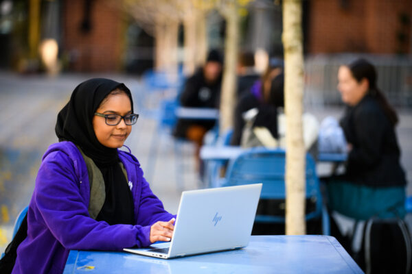 A female student focuses as she works on a laptop while sitting at an outdoor patio table.