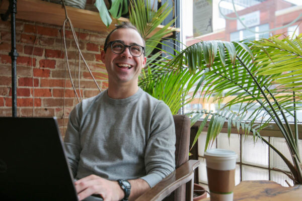 A smiling male student looks up from working on a laptop in a coffee shop.