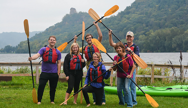 Students and their parents pose together near Lake Winona wearing lifevests and holding kayak paddles.