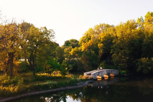 People walk across a small bridge in a wooded park.