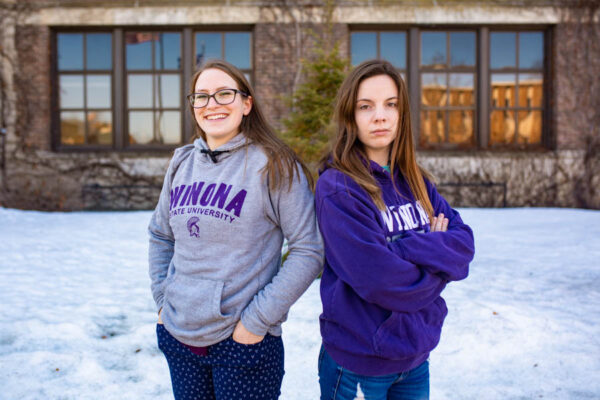 Two female students pose on campus, one looking happy and one looking frustrated.