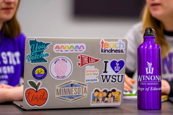 Two students work on a laptop that is covered in stickers about WSU, being a teacher, and Minnesota.