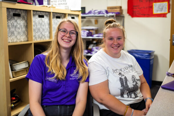 Two student workers smile while sitting behind an office desk.