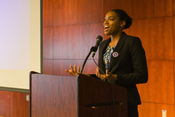 A female speaker presents on stage at a campus event.