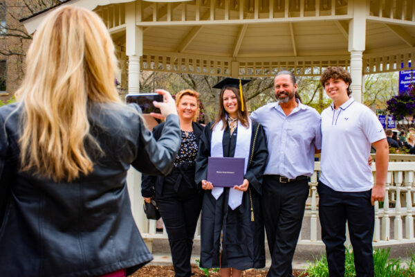 A graduate in cap and gown poses with family for a photo in front of the gazebo on WSU campus.