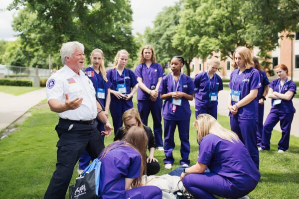 A group of students in purple scrubs watch a nursing demonstration with a mannikin.