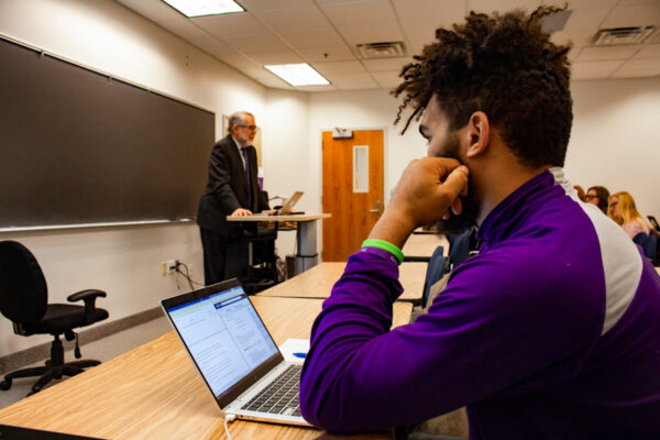 A student takes notes on a laptop while a professor speaks to a class.