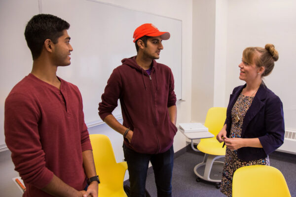 Two students chat with a professor in a classroom.