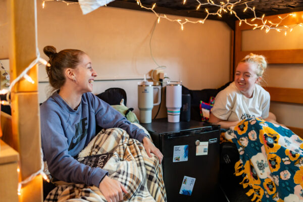 Two female student hang out in a small living space below a lofted bed in a residence hall room.