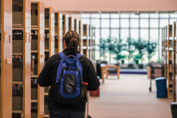 A student wearing a backpack walks past bookshelves in the WSU Library.