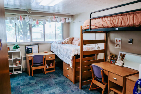 A room in Richards Hall with two lofted beds along one wall, a desk tucked beneath one bed and another desk placed by the windows.