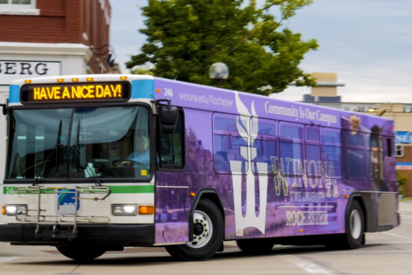 A city bus wrapped in an ad for WSU-Rochester travels down a city street.