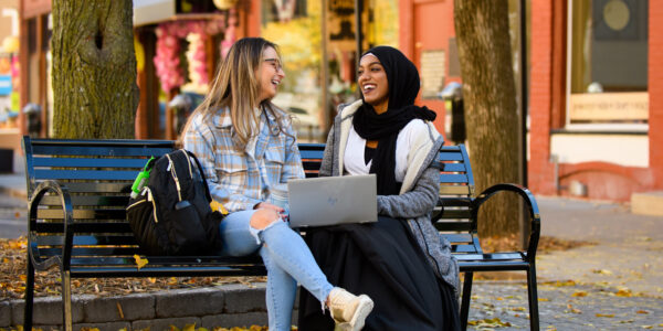 Two students laugh and talk while sitting on a bench on a sidewalk in Rochester, MN.
