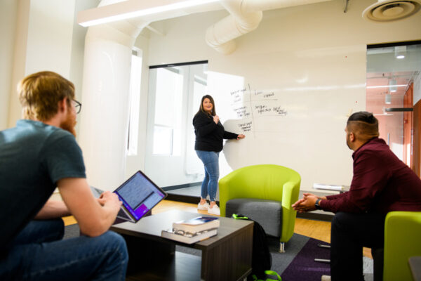 Three students work on a group project using their laptops and a whiteboard in a meeting room at the WSU-Rochester Broadway location.