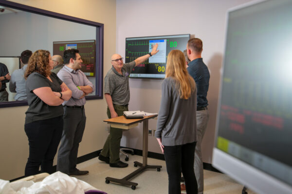 A professor instructs a group of students on reading a chart in a Nursing Simulation lab.