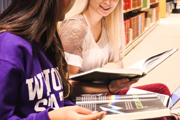 Two students look at books together by shelves in a library.